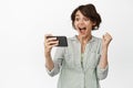 Image of happy brunette girl watching something on mobile phone, cellular 4g internet, celebrating win online, standing Royalty Free Stock Photo