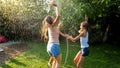 Photo of happy cheerful girls in wet clothes dancing and jumping under water garden hose. Family playing and having fun Royalty Free Stock Photo