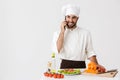 Image of happy chef man in uniform smiling and talking on smartphone while cooking vegetable salad