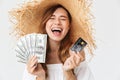 Image of happy rich woman 20s wearing big straw hat laughing while showing credit card and fan of dollar bills, isolated over Royalty Free Stock Photo