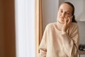 Image of helpless confused woman wearing beige sweater posing near window, talking on cell phone, being not sure what to answer, Royalty Free Stock Photo