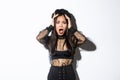 Image of horrified and shocked asian woman in gothic lace dress and wreath looking ambushed, wearing halloween costume