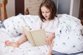 Image of impressed surprised little girl having shocked facial expression, reading fairytale alone, sitting on her bed, having