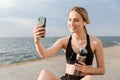 Image of joyful woman taking selfie on cellphone and holding water bottle while sitting on mat near seaside Royalty Free Stock Photo