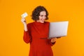 Image of joyous woman using silver laptop and credit card while standing isolated over yellow background