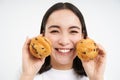 Image of korean woman enjoys eating baked pastry, showing two tasty cupcakes near face and smiles, white background Royalty Free Stock Photo