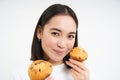 Image of korean woman enjoys eating baked pastry, showing two tasty cupcakes near face and smiles, white background Royalty Free Stock Photo