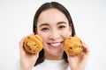 Image of korean woman enjoys eating baked pastry, showing two tasty cupcakes near face and smiles, white background Royalty Free Stock Photo