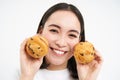 Image of korean woman enjoys eating baked pastry, showing two tasty cupcakes near face and smiles, white background Royalty Free Stock Photo