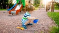 Image of little boy sitting on the playground and puring sand with small plastic spade in colorful bucket. Kid digging Royalty Free Stock Photo