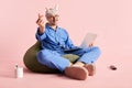 Image of man sitting on bag chair in pajama and funny headband with laptop with love-sign over pastel rose background.