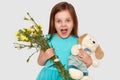 Image of overjoyed blue eyed small child holds her favourite toy and flowers, happy to recieve present on birthday, opens mouth Royalty Free Stock Photo