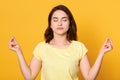 Image of peaceful good looking brunette wearing yellow t shirt, raising hands, having meditation, closing eyes, being alone,