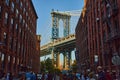 Pillar of Manhattan Bridge in sunlight framed by brick buildings and tourists in street