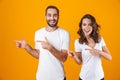 Image of pleased couple man and woman in basic clothing pointing fingers, while standing together isolated over yellow background