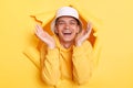 Image of pleased extremely happy man wearing hoodie and panama posing in hole in yellow paper wall, raised arms and being