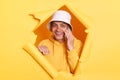 Image of positive optimistic man wearing hoodie and panama posing in hole in yellow paper wall, standing with happy expression and