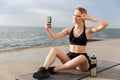 Image of positive woman taking selfie on cellphone and gesturing peace sign while sitting on mat near seaside Royalty Free Stock Photo