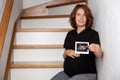 Image of pregnant smiling caucasian curly woman sitting on wooden stairs and posing while showing ultrasound scans on