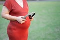 Image of pregnant woman in red maternity clothes standing in garden holding headphone on her baby bump.