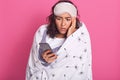 Image of pretty woman dresses white underwear and wrapped blanket with dandelion, being in bedroom with smartphone, looks upset,