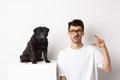 Image of puzzled young man sitting near cute black pug, showing small size and frowning disappointed, white background