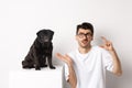 Image of puzzled young man sitting near cute black pug, showing small size and shrugging confused, white background.