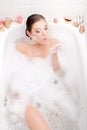 Image of relaxing beautiful young woman lying in a spa bath with foam and shell blowing soap bubbles