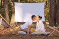 Image of romantic couple sitting in the forest with overhead projector with empty display for advertisement, husband kissing his Royalty Free Stock Photo