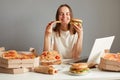 Image of satisfied delighted Caucasian woman eating fast food, sitting at table with lots junk food against gray wall, holding