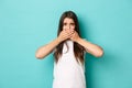 Image of scared brunette girl in white t-shirt, looking anxious and shut mouth with hands, standing over blue background