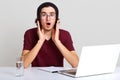 Image of scared young man working on laptop, wearing maroon t shirt and black cap, posing with open mouth and shocked facial