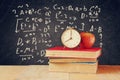 Image of school books on wooden desk, apple and vintage clock over black background with formulas. education concept