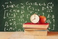 Image of school books on wooden desk, apple and vintage clock over green background with formulas. education concept