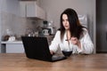 An image of a serious beautiful woman in a bathrobe taking notes and using a laptop while sitting at the table in the Royalty Free Stock Photo