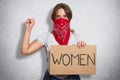 Image of serious bossy militant young feminist raising arm, showing fist, holding sign in one hand, having inscription women on
