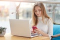 Image of serious busy young girl working online, sitting at cafe, using her laptop and mobile phone simultaneously, having long Royalty Free Stock Photo