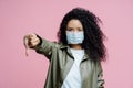 Image of serious curly haired young woman wears medical mask, keeps thumb down, shows dislike gesture, avoids spreading of corona