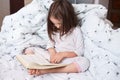 Image of serious preschooler girl reading book in bed, cute kid sitting under blanket with dandelion, darkhaired child reads fairy