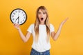 Shocked displeased young blonde woman posing isolated over yellow wall background dressed in white casual t-shirt holding clock