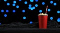 A red cup of popcorn with a red and white striped straw sits on a black surface, with a background of blue bokeh lights. Royalty Free Stock Photo
