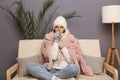 Image of sick ill unhealthy attractive woman wearing cap and winter coat sitting on the sofa in cold flat, feeling unwell, Royalty Free Stock Photo