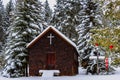 Image of a small rural church sitting amongst snow covered spruce trees, one which has a few red Christmas ornaments on it.