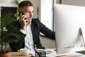 Image of smart businessman talking on cell phone while working on computer in office Royalty Free Stock Photo