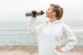 Image of smiling blond woman using earpods and drinking water from bottle while walking near seaside Royalty Free Stock Photo