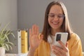 Image of smiling cheerful dark haired young adult woman freelancer using smart phone in office, having video call, saying hello to Royalty Free Stock Photo