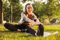 Image of smiling fitness woman 20s wearing headphones working out and stretching legs, while sitting on exercise mat in green park