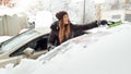 Close up image of smiling girl in jacket trying to clean up snow covered auto by brush. Scraping the windshield and