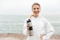 Image of smiling happy woman using earpods and drinking water from bottle while walking near seaside Royalty Free Stock Photo