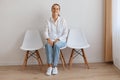 Image of smiling satisfied attractive young adult woman sitting on chair against white wall, wearing shirt and jeans, looking at Royalty Free Stock Photo